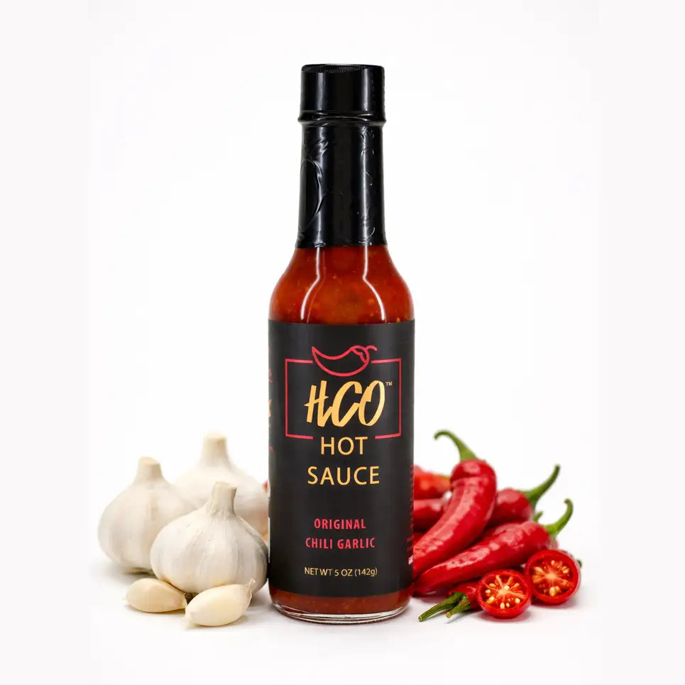 A bottle of HCO Hot Sauce, Original Chili Garlic flavor, displayed on a white background with whole and sliced red chili peppers and garlic bulbs arranged around the base of the bottle.