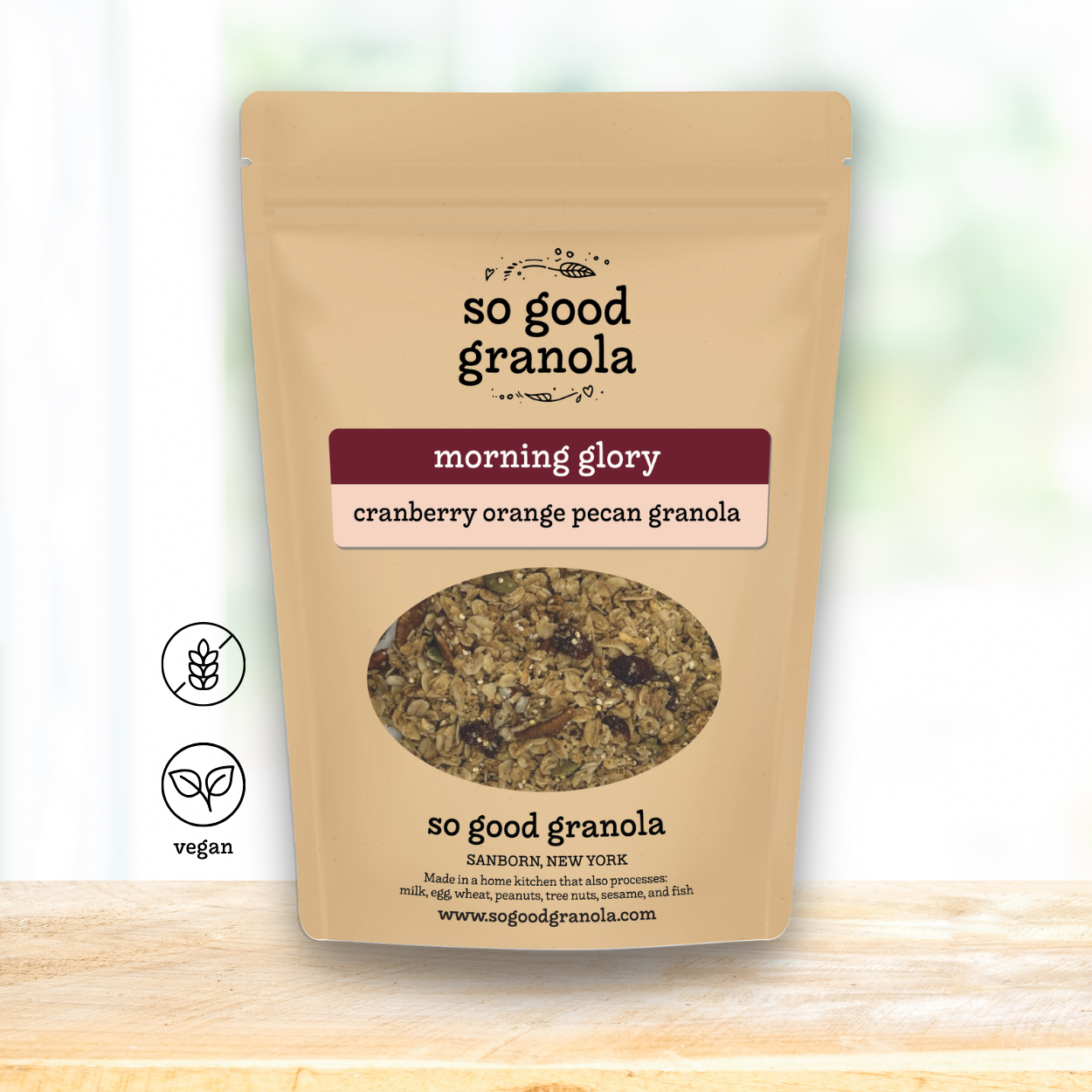 A stand-up pouch of So Good Granola in the 'morning glory' flavor, featuring a kraft paper design with a circular window showing the granola inside. The pouch is displayed on a light wooden surface against a soft, blurred white background with two 'vegan' certification icons on the left.