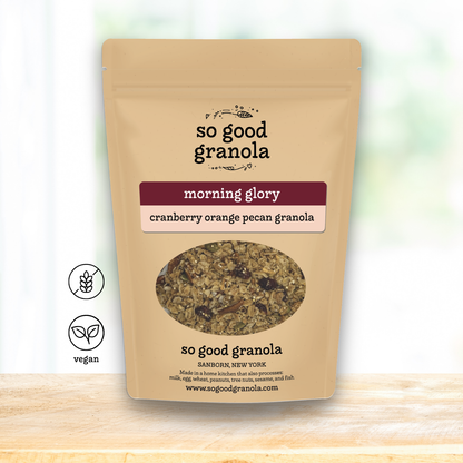 A stand-up pouch of So Good Granola in the 'morning glory' flavor, featuring a kraft paper design with a circular window showing the granola inside. The pouch is displayed on a light wooden surface against a soft, blurred white background with two 'vegan' certification icons on the left.