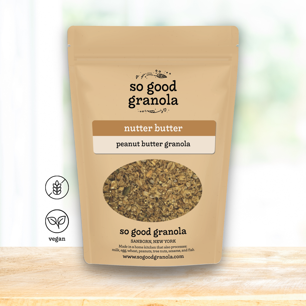 A stand-up pouch of So Good Granola Nutter Butter / Peanut Butter Granola on a light wooden surface with a soft white background. The packaging is beige with brown and white label blocks, a clear oval window showing the granola inside, and two small icons on the left indicating nut butter and vegan.