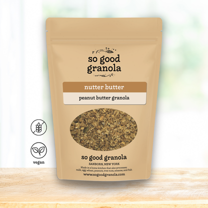A stand-up pouch of So Good Granola Nutter Butter / Peanut Butter Granola on a light wooden surface with a soft white background. The packaging is beige with brown and white label blocks, a clear oval window showing the granola inside, and two small icons on the left indicating nut butter and vegan.
