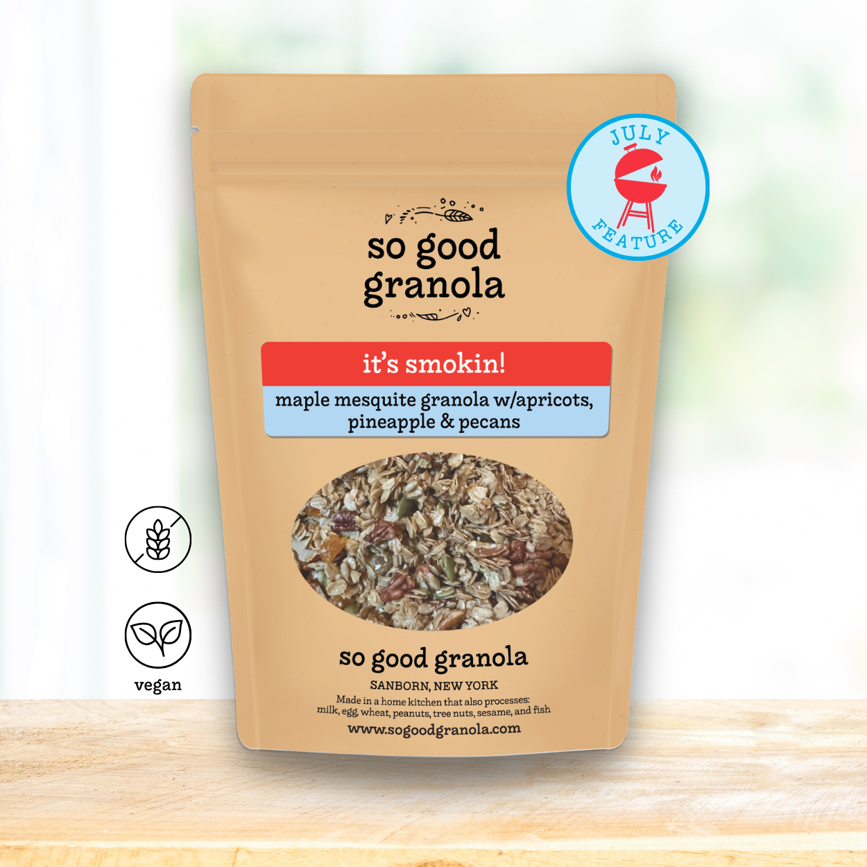 A stand-up pouch of So Good Granola in the 'it's smokin!' flavor, featuring a kraft paper design with a circular window showing the granola inside. The pouch has a blue 'July Feature' badge and is displayed on a light wooden surface against a bright, blurred background.