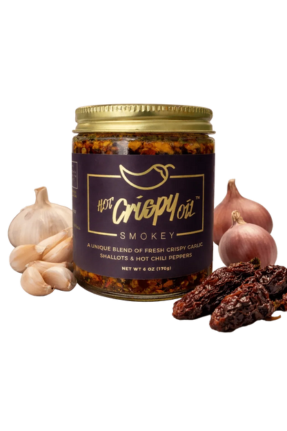 A jar of Hot Crepy Oil Smokey chili oil surrounded by fresh ingredients including garlic cloves, whole garlic bulbs, and dried red chilies on a white background.